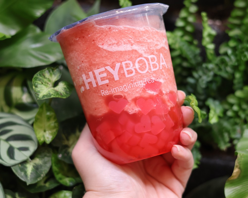 Bright pink HEYBOBA fruit tea held among tropical plants
